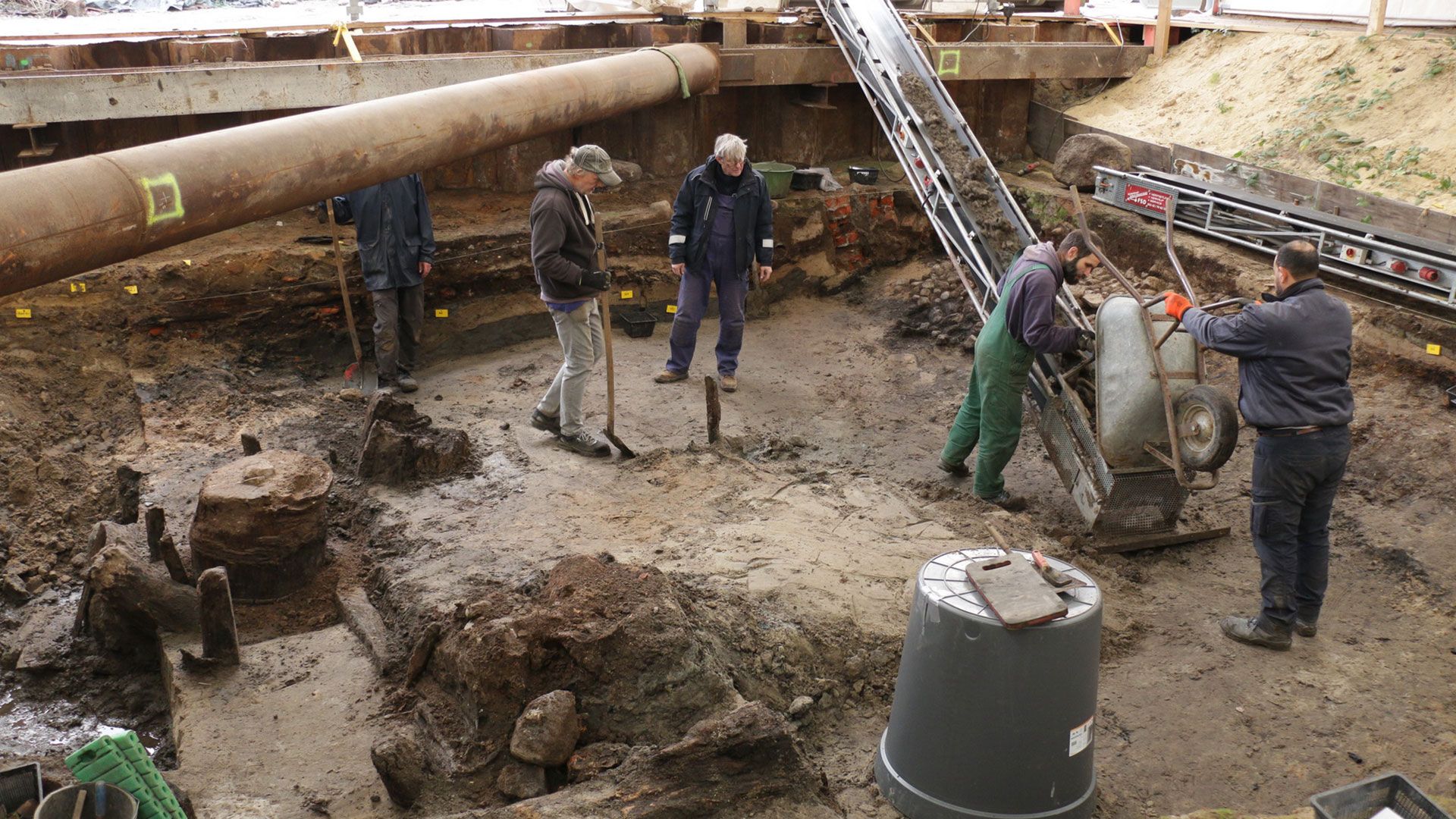 Blick auf das Fußbodenniveau von 1536. Links im Bild ist deutlich ein gut erhaltenes Holzfass zu erkennen, rechts unter dem Förderband die Reste eines Steinpflasters. | Foto: Archäologisches Museum Hamburg.