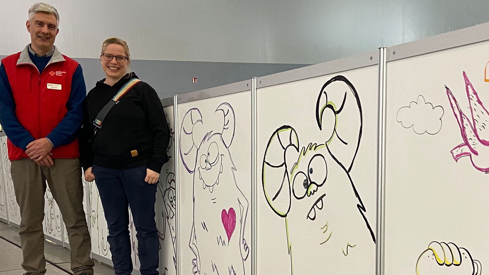 Einrichtungsleiter des DRK-Hamburg-Harburg Robert Jesse und Künstlerin Melanie Groger vor der Sichtschutzwand mit den schielenden Monstern, gemalt gemeinsam mit den Kindern der Unterkunft. | Foto: DRK